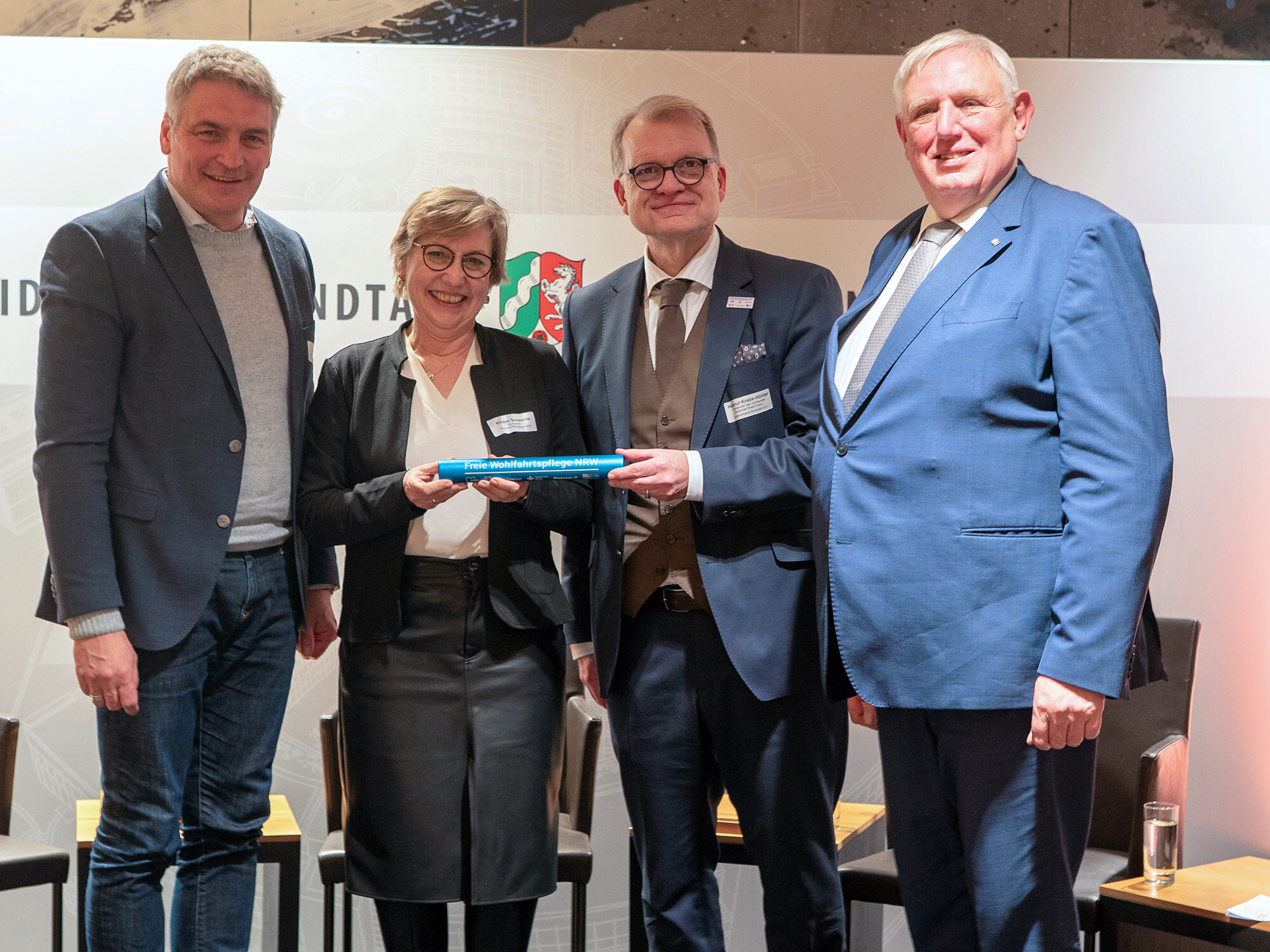 Vier Personen in formeller Kleidung stehen nebeneinander und präsentieren ein blaues Objekt mit der Aufschrift "Freie Wohlfahrtspflege NRW".